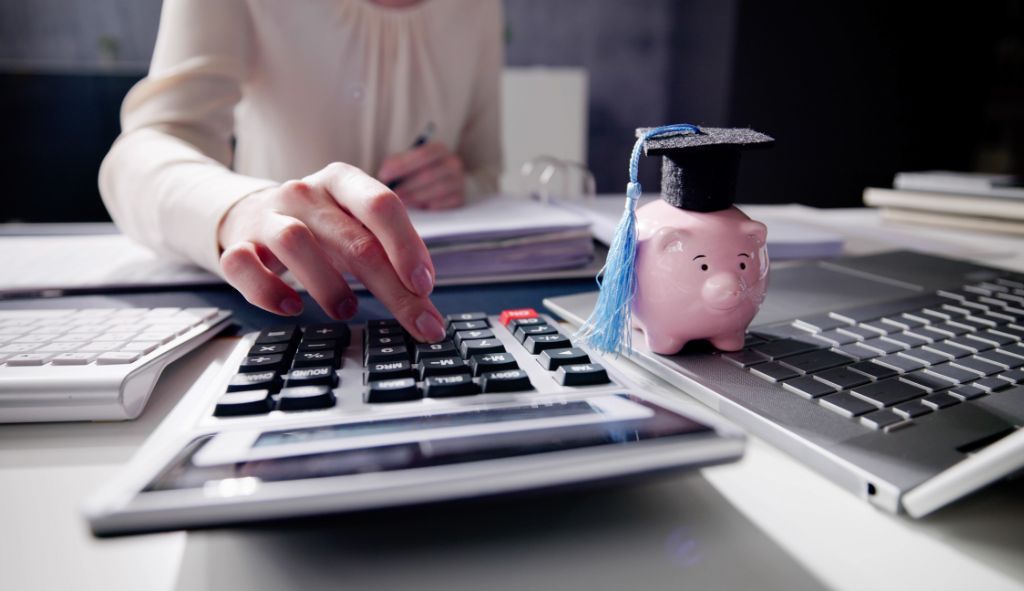 eine Frauenhand die auf einem Taschenrechner tippt. Ein Laptop und ein Sparschwein